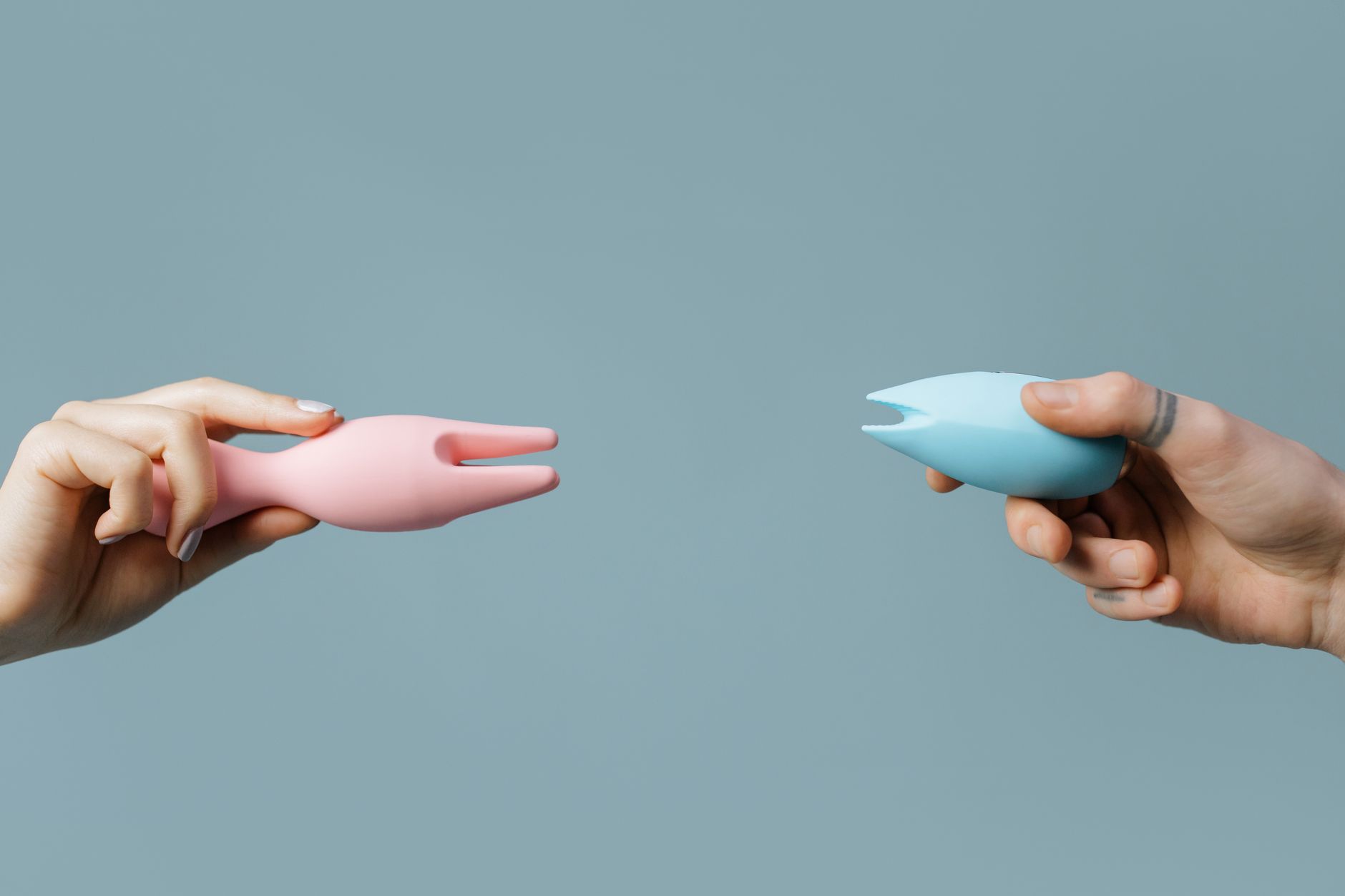 Two hands holding pink and blue silicone vibrators against a pastel background, symbolizing shared intimacy and connection.