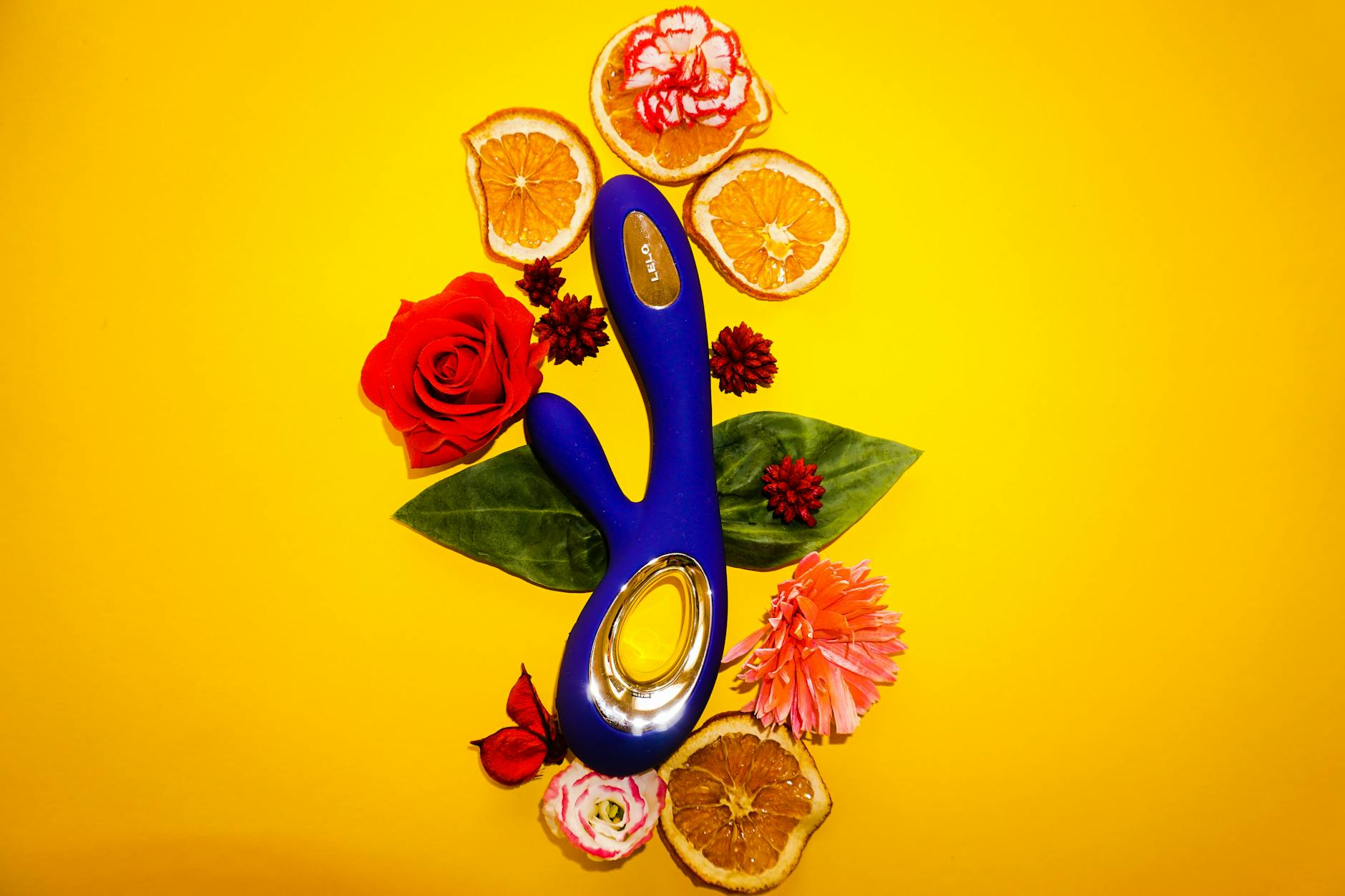 Ein blauer Vibrator umgeben von Blumen und Orangenscheiben auf gelbem Untergrund.